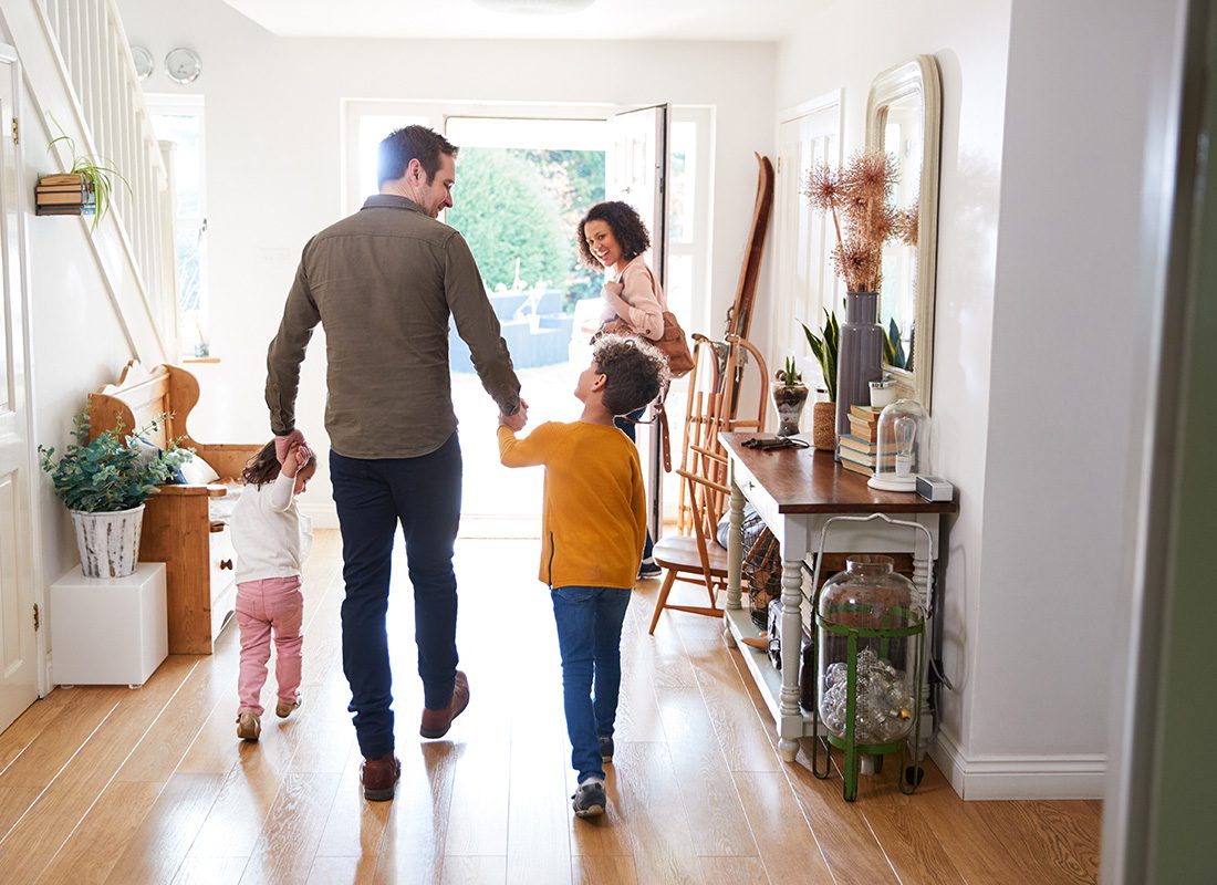 Personal Insurance - Rear View of a Father Holding Hands with his Two Children Walking Toward the Front Door of Their Home While the Mother Holds Open the Door